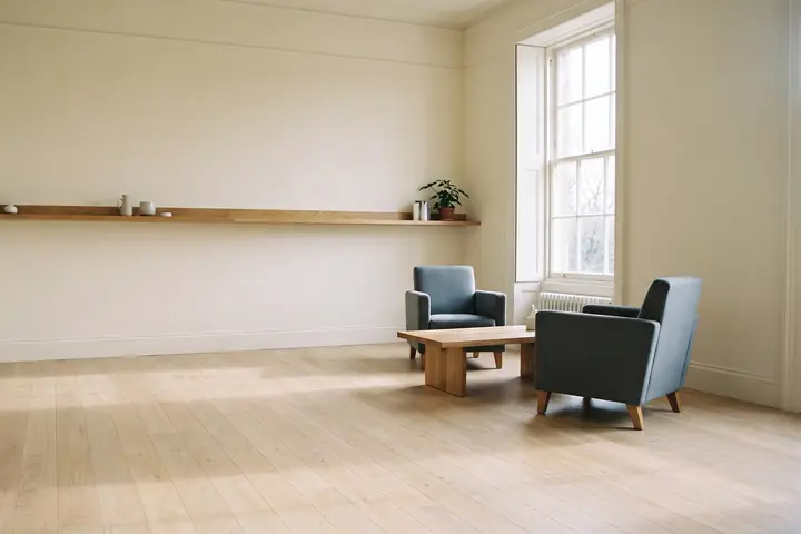 An empty, light-filled room with two armchairs and a long shelf along a plain wall.