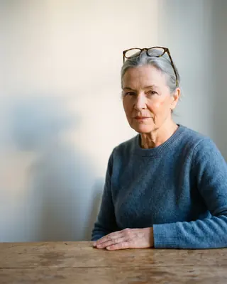 A woman in her sixties with grey hair and glasses pushed up, sitting at a bare wooden table in a plain, bright room.